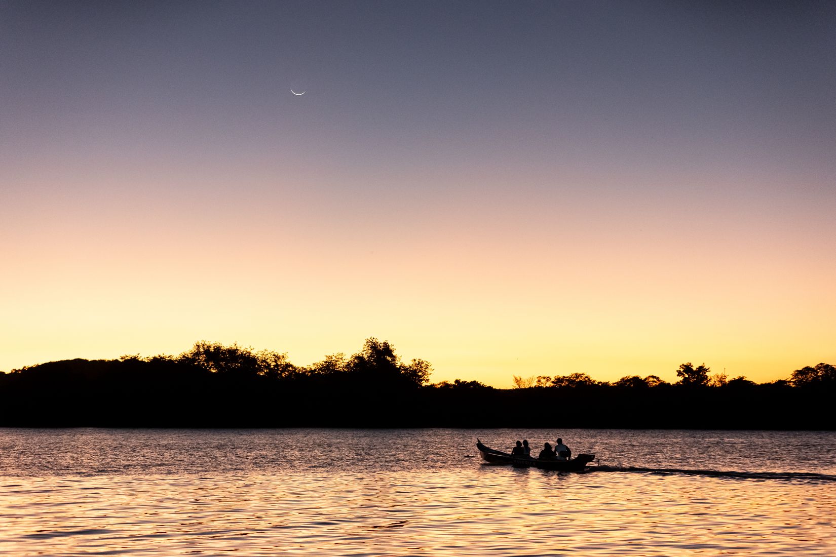 Barco navegando ao pôr do sol em rio brasileiro, com a lua crescente no céu
